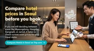A professional and inviting horizontal banner featuring a woman sitting at a modern hotel lobby desk in Seoul, using a laptop to compare hotel prices. On the left, a dark overlay with white and gold text reads: "Compare hotel prices in Seoul before you book. If you are still deciding between areas like Myeongdong, Hongdae, Gangnam, or Jamsil, it helps to check current room prices for your travel dates." Below the text, there is a prominent teal button that says "Compare Hotels in Seoul on Trip.com →". A small map of Seoul with highlighted tourist districts is visible in the background, and a friendly hotel staff member is assisting her, creating a sense of reliable travel planning.