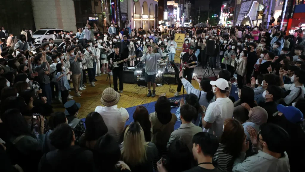 A vibrant nighttime photograph capturing a high-energy live busking performance in the bustling Hongdae district of Seoul, South Korea. A massive crowd of young people, densely packing the square with some still wearing masks, surrounds a three-piece live band (guitarist, vocalist, and bassist with their instruments) performing live on a textured yellow-tiled street. All elements, including individual faces in the crowd, the band's detailed expressions and equipment, and the bright banner reading 'HONGDAE LIVE PERFORMANCE | BUSKING NIGHTS | #HONGIKUNIV' and all surrounding building signs (in Korean and English), are incredibly sharp and legible. The complex city grid with multiple buildings, all brightly illuminated with specific names, is visible in the distance under a clear night sky. The scene is illuminated by diverse but defined light sources, creating a lively atmosphere. Every single detail and facial expression is defined, making it look like a professional, high-end photograph. The ground is defined by detailed pavers and clear textures. All faces are clear and distinct. The buildings in the far distance are distinct, showing a dense city grid