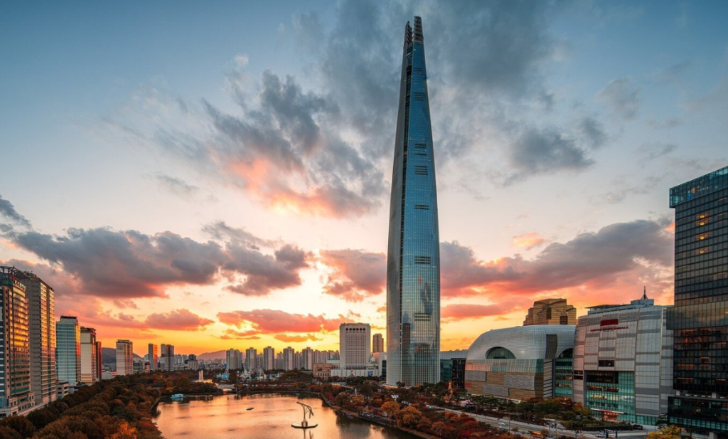 A stunning sunset view of the Lotte World Tower, the tallest skyscraper in South Korea, piercing through a dramatic orange and blue sky. The photo captures the reflection of the sunset in Seokchon Lake in the foreground, with the modern city skyline of Songpa-gu and Lotte World Mall visible at the base. This image showcases the "Sky-High Luxury" experience of staying at SIGNIEL SEOUL, offering a panoramic view of the city's modern architecture and natural beauty.