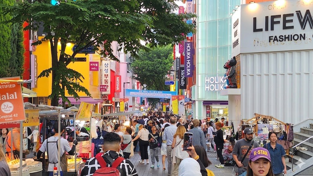 A bustling daytime scene at Myeongdong shopping street in Seoul, South Korea, crowded with international tourists and locals. The street is lined with vibrant shops featuring colorful signs for brands like Holika Holika and Missha. Street food stalls with various snacks and Halal-certified options are visible in the foreground, creating a lively atmosphere characteristic of Seoul's most famous shopping district.