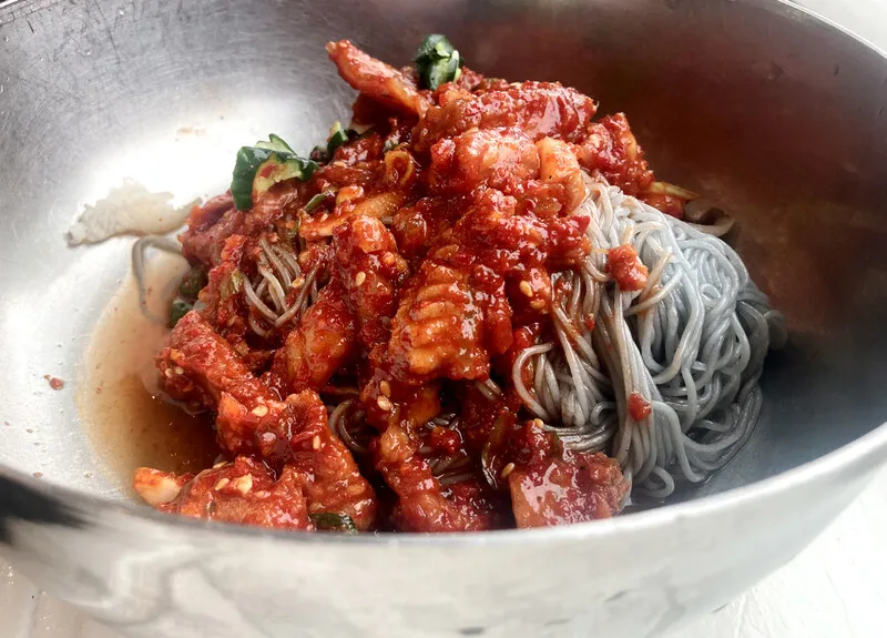 Spicy Hamheung naengmyeon in a metal bowl, with chewy gray buckwheat noodles topped with red chili sauce, sliced raw fish, and vegetables.